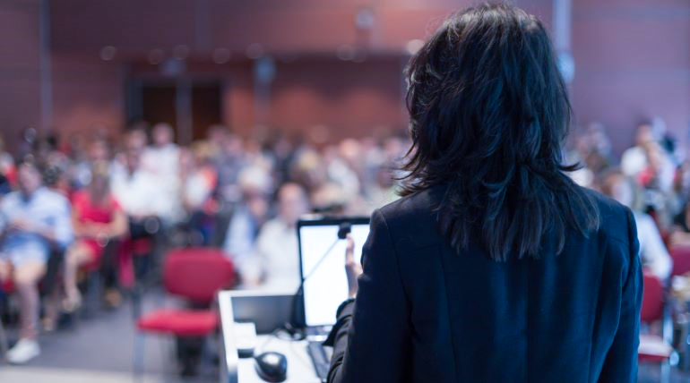 Kvinna står vid en mikrofon framför människor som sitter i en konferenssal. 