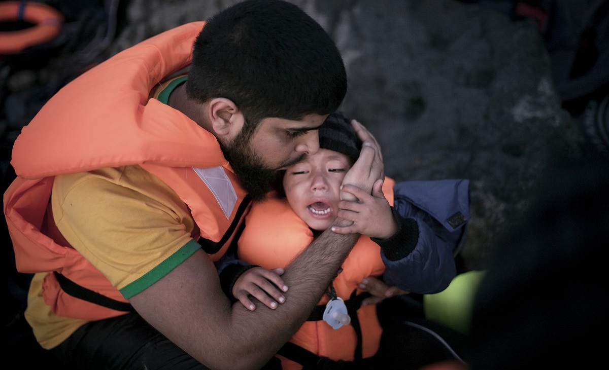 En far kramar om sin gråtande son. Båda är iförda flytvästar och en livräddningsboj ligger en bit bort.